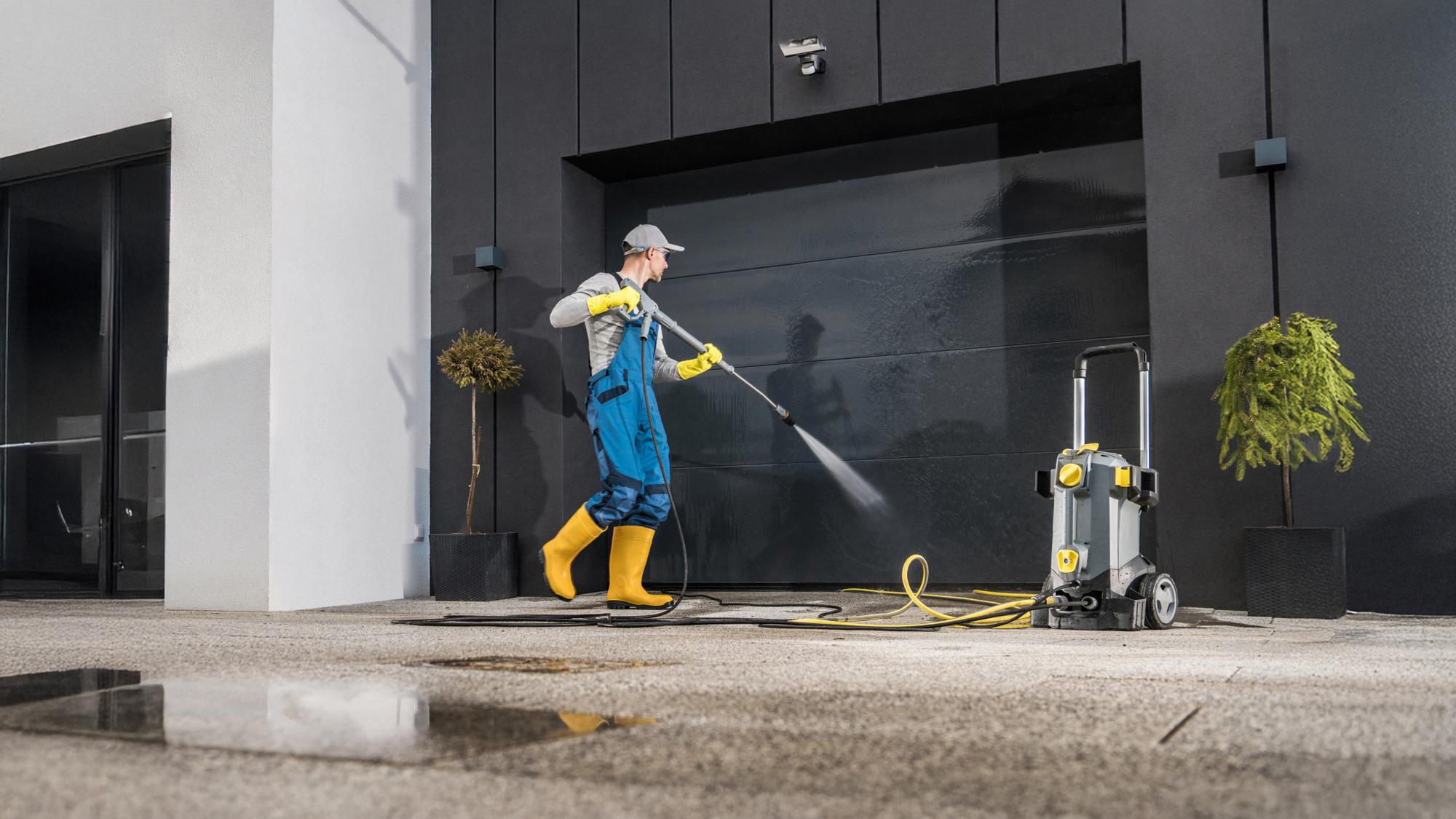 A person in blue overalls and yellow rubber boots is using a pressure washer to clean a dark garage door in a modern setting, with a potted plant nearby and wet pavement.